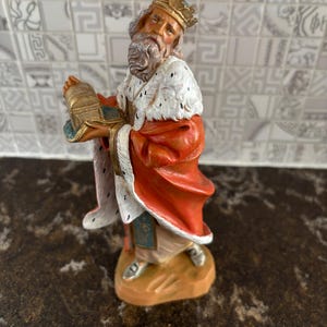 May include: A figurine of a king, part of a nativity scene. The king wears a gold crown, a red robe with white fur trim, and holds a gold chest. The figurine is on a brown base and has a detailed, aged appearance.