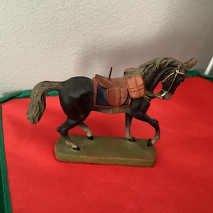 May include: A figurine of a black horse with a saddle and saddlebags. The horse stands on a green base, against a red backdrop. The horse's mane and tail are dark, and the saddle is brown with blue detailing. A decorative item.