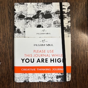 May include: A journal with a black and white abstract design and the text "PILGRIM SOUL". The journal has a black elastic band and a white label with the text "PLEASE USE THIS JOURNAL WHILE YOU ARE HIGH".