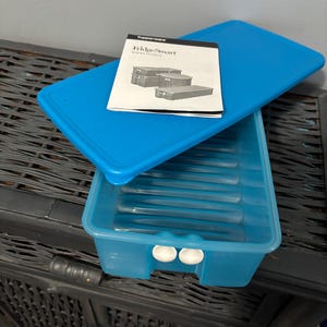 May include: A blue Tupperware FridgeSmart container with a matching lid and instruction manual. The container has a ridged interior and white handles. The manual displays images of the containers. The lid is rectangular.