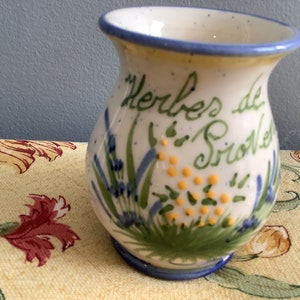 Könnte beinhalten: Eine kleine, weiße Keramikvase mit blauem Rand und einem Blumendesign. Die Vase ist mit grünen Blättern, blauen Blumen und gelben Punkten verziert. Der Text "Herbes de Provence" ist in Kursivschrift auf der Vase geschrieben.