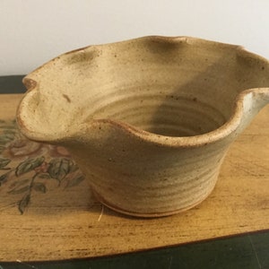 Op de afbeelding: Een handgemaakte, beige keramische kom met een gegolfde rand. De kom heeft een textuuroppervlak en een brede opening. De binnenkant is glad en de buitenkant heeft horizontale ribbels. De kom wordt waarschijnlijk gebruikt om te serveren of tentoon te stellen.