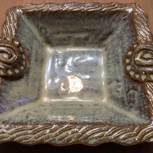 May include: Square ceramic dish with a light green and brown glaze. The rim has a textured, wavy design, and each corner features a decorative spiral element. A handmade pottery piece.