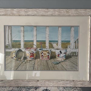 May include: Framed print of beach scene with Mickey Mouse and Donald Duck buckets. The artwork depicts a wooden deck with a view of the ocean and sand dunes. The frame is white with a textured finish.