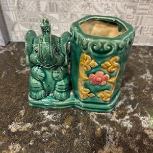 Puede incluir: Una maceta de cerámica verde con una figura de elefante y un recipiente a juego. El elefante tiene la trompa levantada y el recipiente está decorado con detalles florales dorados y rosas. La maceta es un artículo decorativo.