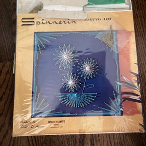 May include: A vintage string art kit featuring a blue background with a design of three white flowers in a blue bowl. The kit includes a pre-printed canvas, string, and nails. The kit is labeled "Spinnerin String Art" and "Bowl of Flowers ST 913".