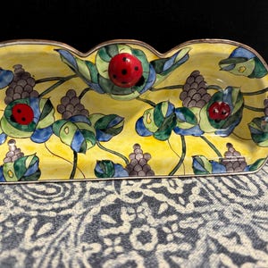 Può includere: Un piatto in ceramica giallo con un design stravagante con coccinelle, uva e foglie. Il piatto ha un bordo smerlato ed è decorato con accenti verdi, blu e viola. Le coccinelle sono rosse con pois neri.