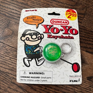 Może przedstawiać: Brelok do kluczy Yo-Yo Duncan na białej i szarej karcie. Zielony yo-yo ma srebrny brelok i czerwony akcent. Karta zawiera postać z kreskówki i tekst "Really Works! High Performance". Pozycja #551-0.