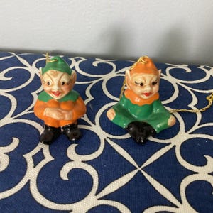 May include: Two ceramic elf ornaments with green hats and orange and green clothing. The elves are sitting with their legs crossed and have black shoes. They have painted faces with red lips and black eyes.