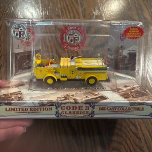 May include: A yellow die-cast model of a Los Angeles City Fire Department fire truck, presented in a clear display case. The packaging features the Code 3 Collectibles logo and the text "Serial Number Certificate Inside Base." It is a Code 3 Classics limited edition.