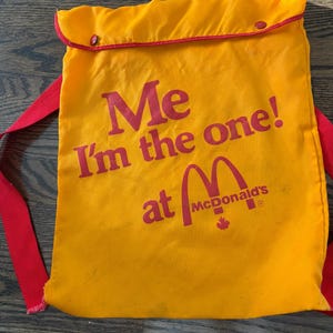 Op de afbeelding: Gele rugzak met trekkoord, rode rand en bandjes. De tas heeft de rode tekst "Me I'm the one! at McDonald's". Het McDonald's-logo is ook zichtbaar.