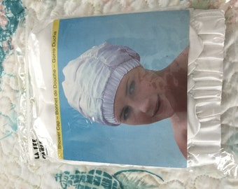 May include: A white shower cap in a clear plastic package. The cap is made of a silky material and has a gathered elastic band. The packaging includes the text "Shower Cap - Bonnet de Douche - Gorro Ducha".