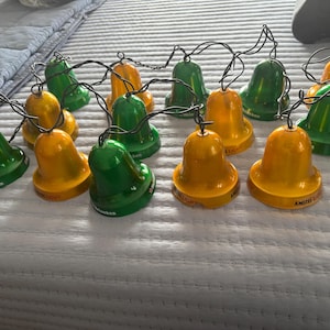 May include: String of bell-shaped string lights in green and yellow. The lights are made from bottle caps and have black wires. The lights are arranged on a light blue surface.