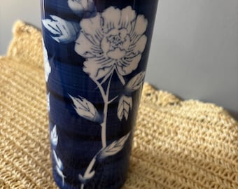 May include: A cylindrical ceramic vase featuring a deep navy blue background with horizontal brushstroke textures. The surface is decorated with a white floral pattern depicting a large blooming flower and leaves extending along the length of the vessel.