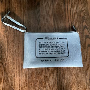 May include: A light blue Coach New York zip-top pouch with a silver-tone wrist strap. The pouch features a black rectangular text box with the text "THIS IS A COACH BAG. THE STORYPATCH IS OUR BADGE OF AUTHENTICITY..." and the numbers "M1632-F36658".