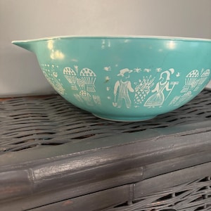 以下が含まれることがあります： 白縁と注ぎ口が付いたターコイズ色のミキシングボウル。ボウルには、人、木、バスケットの白い様式化されたデザインが施されています。ボウルは濃い灰色の籐の表面に置かれています。