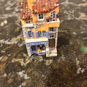 Puede incluir: Una maqueta en miniatura detallada de un edificio europeo. La estructura presenta un tejado de tejas rojas, paredes amarillas y detalles de piedra. Las ventanas tienen contraventanas azules y la fachada muestra el letrero "La vende en Provence".