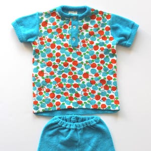 May include: A vintage two-piece terrycloth outfit. The top is a short-sleeved shirt with a blue collar and button placket. The shirt features a pattern of red and blue circles on a white background. Matching blue shorts complete the set.