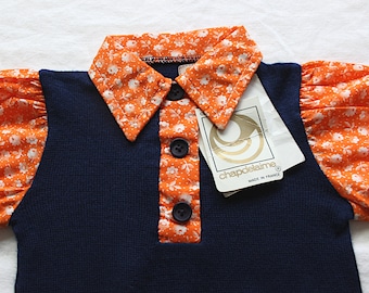Könnte beinhalten: Ein marineblaues Strickhemd mit einem weißen und orangefarbenen Blumenmuster am Kragen und an den Ärmeln. Das Hemd hat einen Knopfverschluss und ein Etikett mit der Aufschrift "chapdelaine made in france".