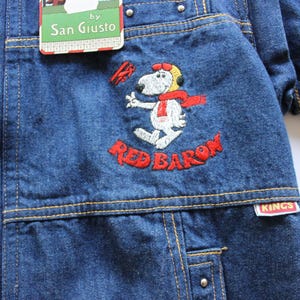 以下が含まれることがあります： スヌーピーがレッドバロンとして刺繍された青いデニムジャケット。 ジャケットは黄色のステッチ、金属リベット、"San Giusto"のタグが特徴です。 "Red Baron"の文字は赤で刺繍されています。 "Kings"のタグも表示されています。