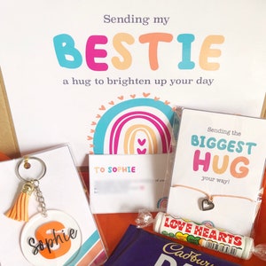 May include: A gift box with a variety of items, including a keychain with the name "Sophie", a candle, a chocolate bar, and a card that says "Sending my BESTIE". The box has a rainbow theme.