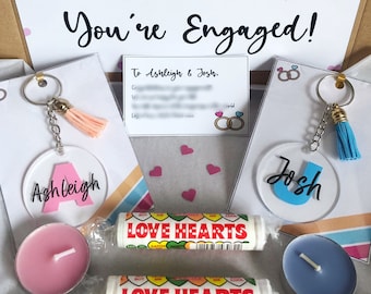 May include: An engagement gift box with a card that says "You're Engaged!" and two keychains with names. The box includes pink and blue candles, and two rolls of Love Hearts candy. Pink hearts are scattered throughout.