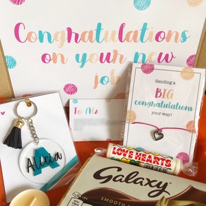 K&ouml;nnte beinhalten: Eine Geschenkbox gef&uuml;llt mit Artikeln zur Feier eines neuen Jobs. Die Box enth&auml;lt eine Tafel Schokolade, eine Karte mit dem Text "Sending a BIG congratulations your way!" und einen Schl&uuml;sselanh&auml;nger mit einem schwarzen Quasten und einem klaren Acryl-Anh&auml;nger mit dem Namen "Alicia" darauf. Die Box enth&auml;lt au&szlig;erdem eine kleine Kerze und ein Armband mit einem Herz-Anh&auml;nger.