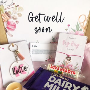 Könnte beinhalten: Geschenk-Korb zum schnelleren Gesundwerden mit einem personalisierten Schlüsselanhänger in Rosa und Weiß, einem rosa und weißen Armband mit einem Nähschere-Anhänger, einer rosa und weißen Karte mit dem Text "Sending a Big Hug your way!" einer rosa und weißen Karte mit dem Text "To Katie", einer kleinen Kerze, einer Cadbury Dairy Milk Schokoladentafel und einer Rolle Love Hearts Bonbons.