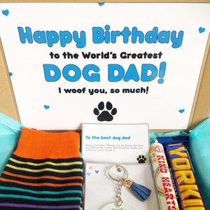 Könnte beinhalten: Eine Geschenkbox gefüllt mit Geburtstagsgeschenken für einen Hundepapa. Die Box enthält ein Paar orangefarbene, schwarze und mehrfarbige gestreifte Socken, einen grünen Pfotenabdruck-Schlüsselanhänger mit dem Namen "Steve" darauf, einen Schokoriegel und eine Karte mit der Aufschrift "Happy Birthday to the World's Greatest Dog Dad! I woof you, so much!"
