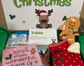 Può includere: Una scatola regalo contenente un biglietto con il testo 'ITS BEGINNING TO LOOK ALOT LIKE Christmas' e un disegno di renna. Include un biglietto 'To Katie', una sagoma di omino di pan di zenzero, un sottobicchiere rosa, un calzino rosso e popcorn.