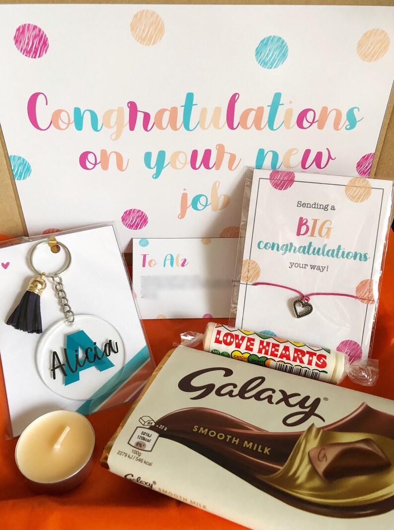 K&ouml;nnte beinhalten: Eine Geschenkbox gef&uuml;llt mit Artikeln zur Feier eines neuen Jobs. Die Box enth&auml;lt eine Tafel Schokolade, eine Karte mit dem Text "Sending a BIG congratulations your way!" und einen Schl&uuml;sselanh&auml;nger mit einem schwarzen Quasten und einem klaren Acryl-Anh&auml;nger mit dem Namen "Alicia" darauf. Die Box enth&auml;lt au&szlig;erdem eine kleine Kerze und ein Armband mit einem Herz-Anh&auml;nger.