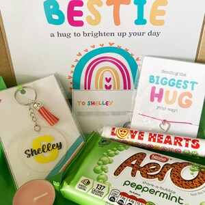 May include: A gift box with a variety of items, including a keychain with a tassel, a candle, a bracelet, and a candy bar. The box also contains a card that says "Sending my BESTIE a hug to brighten up your day."