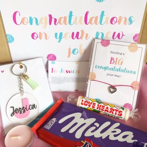 K&ouml;nnte beinhalten: Eine Geschenkbox gef&uuml;llt mit Artikeln zur Feier eines neuen Jobs. Die Box enth&auml;lt eine rosa und wei&szlig;e Karte mit dem Text "Herzlichen Gl&uuml;ckwunsch zu deinem neuen Job", eine rosa und wei&szlig;e Karte mit dem Text "Sende dir ein gro&szlig;es Gl&uuml;ckwunsch auf deinem Weg!" einen rosa Schl&uuml;sselanh&auml;nger mit einem Quasten, eine rosa Kerze, eine Schokoladentafel, ein rosa Armband mit einem Herz-Anh&auml;nger und eine rosa und wei&szlig;e Karte mit dem Text "An Jessica".