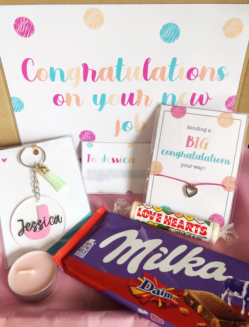 K&ouml;nnte beinhalten: Eine Geschenkbox gef&uuml;llt mit Artikeln zur Feier eines neuen Jobs. Die Box enth&auml;lt eine rosa und wei&szlig;e Karte mit dem Text "Herzlichen Gl&uuml;ckwunsch zu deinem neuen Job", eine rosa und wei&szlig;e Karte mit dem Text "Sende dir ein gro&szlig;es Gl&uuml;ckwunsch auf deinem Weg!" einen rosa Schl&uuml;sselanh&auml;nger mit einem Quasten, eine rosa Kerze, eine Schokoladentafel, ein rosa Armband mit einem Herz-Anh&auml;nger und eine rosa und wei&szlig;e Karte mit dem Text "An Jessica".
