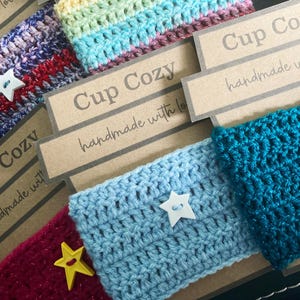 Könnte beinhalten: Eine Sammlung von gehäkelten Tassenmanschetten in verschiedenen Farben wie Blaugrün, Hellblau, Magenta und mehrfarbigem Garn. Jede Manschette hat einen sternförmigen Knopf und ein braunes Papieretikett mit der Aufschrift Cup Cozy handmade with love.