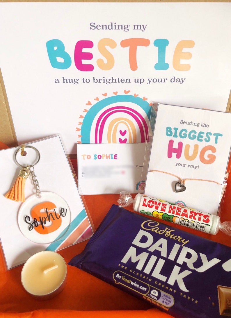 May include: A gift box with a variety of items, including a keychain with the name "Sophie", a candle, a chocolate bar, and a card that says "Sending my BESTIE". The box has a rainbow theme.