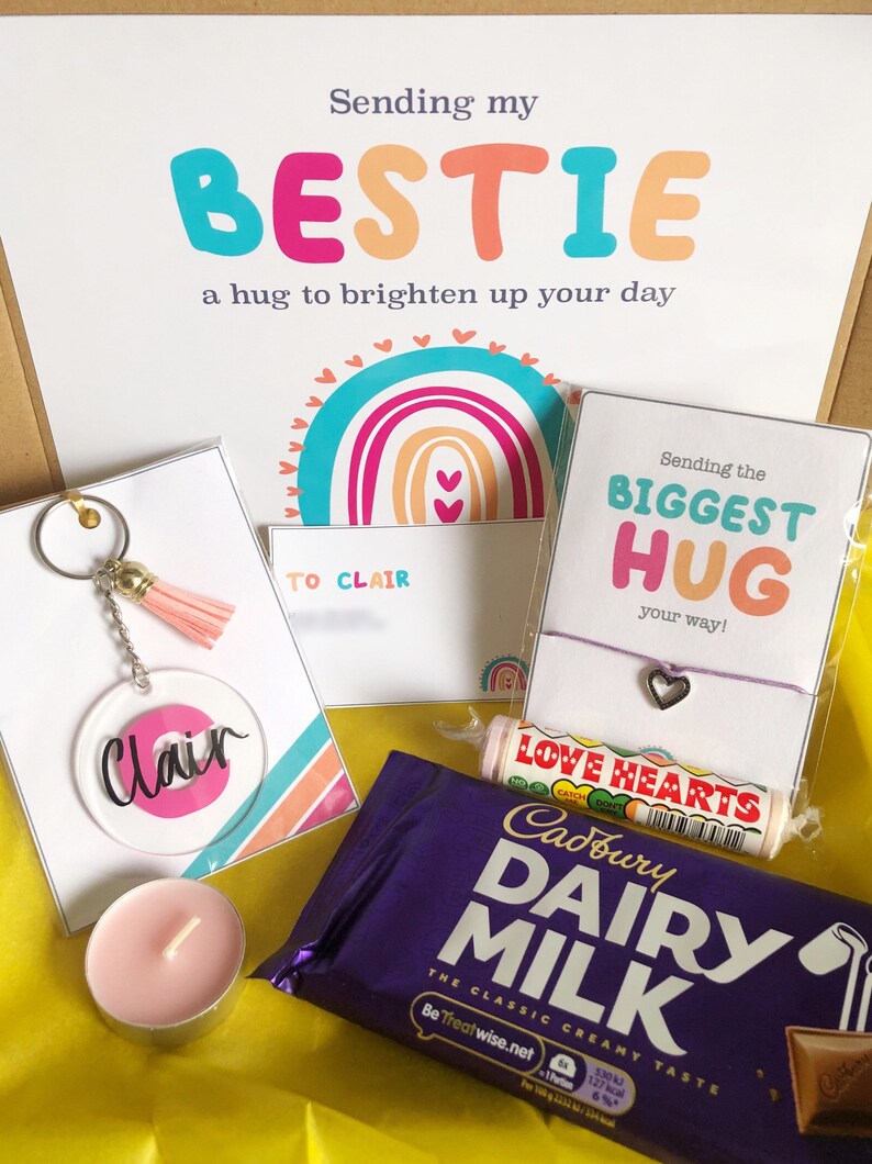 May include: A gift box with a variety of items, including a keychain, bracelet, candy, chocolate bar, and a candle. The box contains a card that says "Sending my BESTIE a hug to brighten up your day". The keychain has a pink tassel.