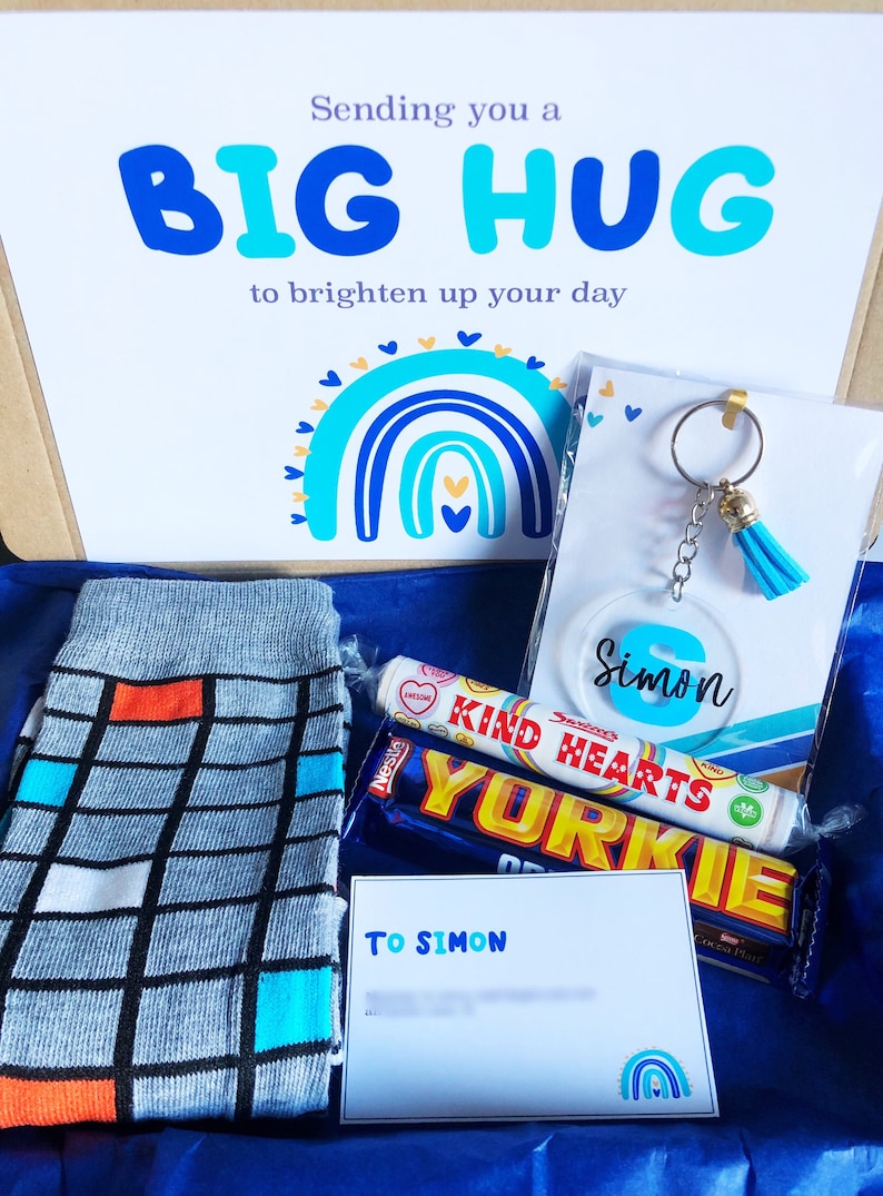以下が含まれることがあります： 友だちへのギフトボックス。ボックスには、青とオレンジの靴下、チョコレートバー、ハート型のキャンディー、青いタッセル付きのキーホルダー、そして「To Simon」と書かれたカードが入っています。