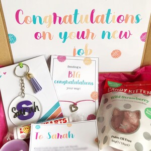 K&ouml;nnte beinhalten: Eine rosa Geschenkbox gef&uuml;llt mit verschiedenen Artikeln zur Feier eines neuen Jobs. Die Box enth&auml;lt einen personalisierten Schl&uuml;sselanh&auml;nger, ein Armband, eine Kerze, einen Lippenbalsam, eine Karte mit dem Text "Herzlichen Gl&uuml;ckwunsch zu deinem neuen Job" und eine T&uuml;te S&uuml;&szlig;igkeiten.
