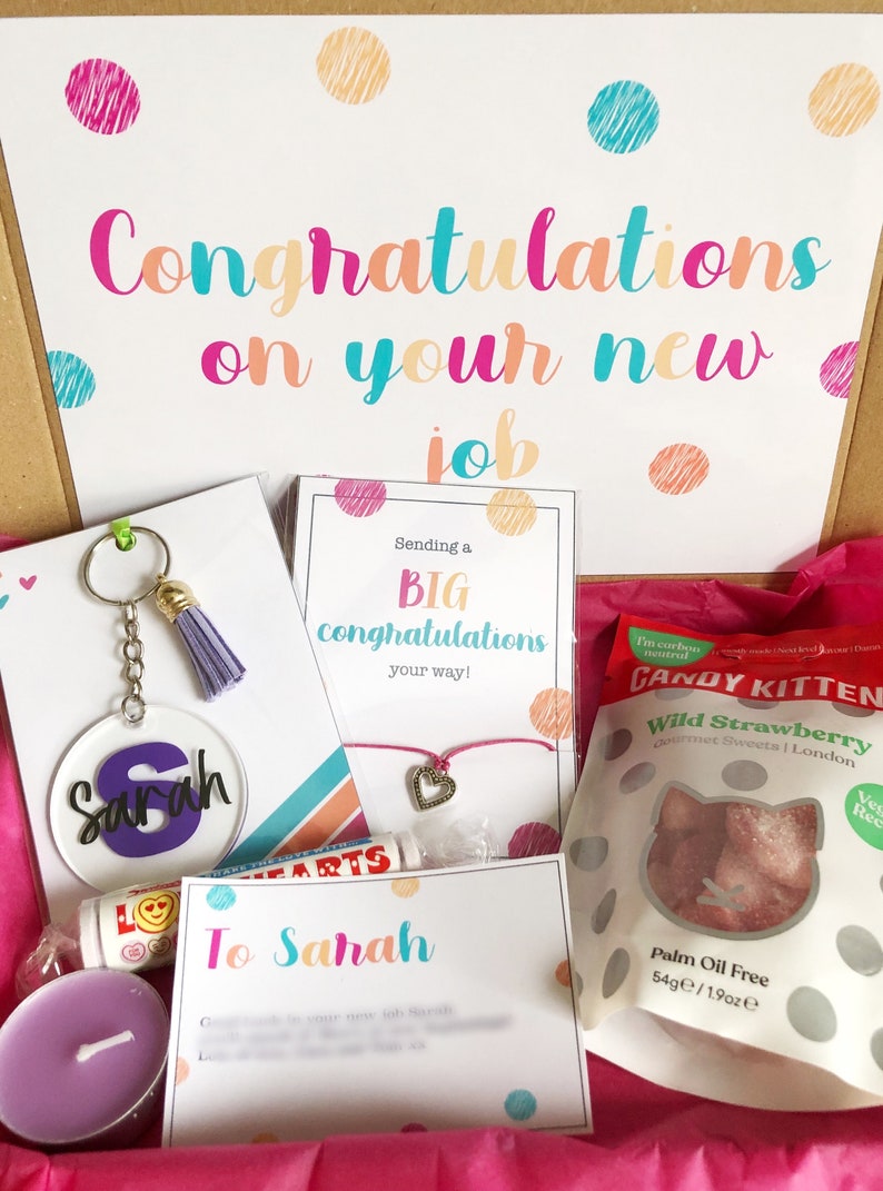 K&ouml;nnte beinhalten: Eine rosa Geschenkbox gef&uuml;llt mit verschiedenen Artikeln zur Feier eines neuen Jobs. Die Box enth&auml;lt einen personalisierten Schl&uuml;sselanh&auml;nger, ein Armband, eine Kerze, einen Lippenbalsam, eine Karte mit dem Text "Herzlichen Gl&uuml;ckwunsch zu deinem neuen Job" und eine T&uuml;te S&uuml;&szlig;igkeiten.