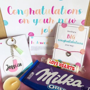 K&ouml;nnte beinhalten: Eine Geschenkbox gef&uuml;llt mit Artikeln zur Feier eines neuen Jobs. Die Box enth&auml;lt eine Karte mit dem Text "Herzlichen Gl&uuml;ckwunsch zu deinem neuen Job", eine Tafel Schokolade, einen Schl&uuml;sselanh&auml;nger, eine Kerze, ein Armband und eine Karte mit dem Text "Wir senden dir ganz herzliche Gl&uuml;ckw&uuml;nsche zu deinem neuen Job!"