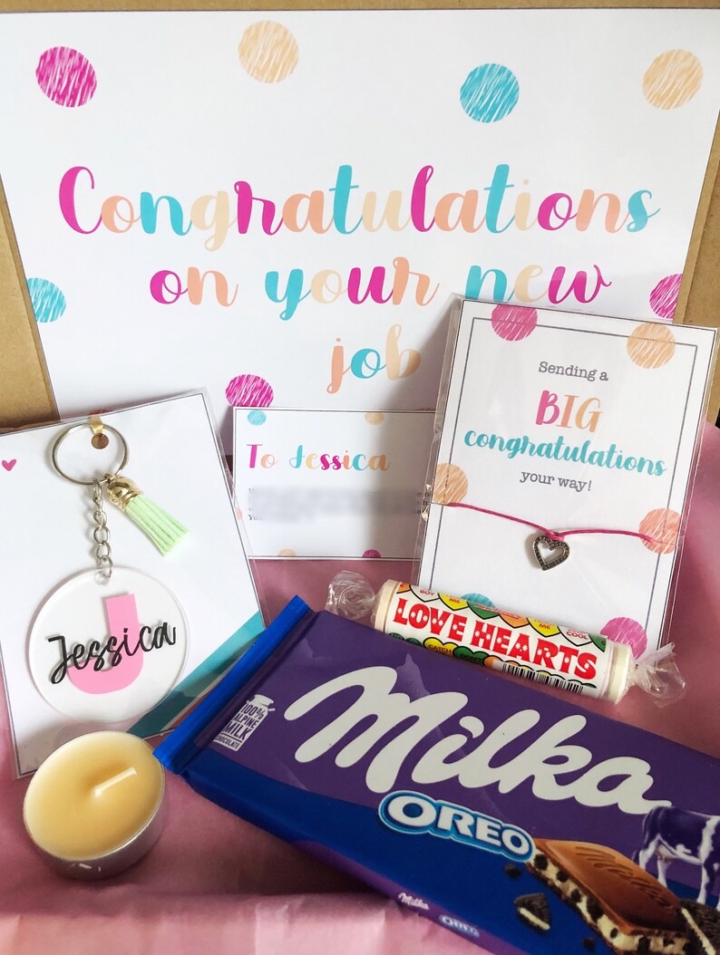 K&ouml;nnte beinhalten: Eine Geschenkbox gef&uuml;llt mit Artikeln zur Feier eines neuen Jobs. Die Box enth&auml;lt eine Karte mit dem Text "Herzlichen Gl&uuml;ckwunsch zu deinem neuen Job", eine Tafel Schokolade, einen Schl&uuml;sselanh&auml;nger, eine Kerze, ein Armband und eine Karte mit dem Text "Wir senden dir ganz herzliche Gl&uuml;ckw&uuml;nsche zu deinem neuen Job!"