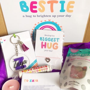 May include: A gift box filled with a variety of items, including a keychain with the name "Zaya", a lip balm, a heart charm bracelet, and a bag of sour watermelon candies. The box also contains a card that says "Sending my BESTIE a hug to brighten up your day".