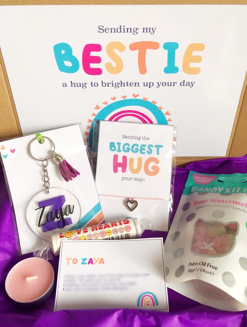 May include: A gift box filled with a variety of items, including a keychain with the name "Zaya", a lip balm, a heart charm bracelet, and a bag of sour watermelon candies. The box also contains a card that says "Sending my BESTIE a hug to brighten up your day".