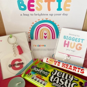 May include: A gift box with a variety of items, including a keychain with a pink tassel, a card with the name "Gemma", a candy bar, a candle, and a bracelet. The box includes a card that says "Sending my BESTIE a hug to brighten up your day".