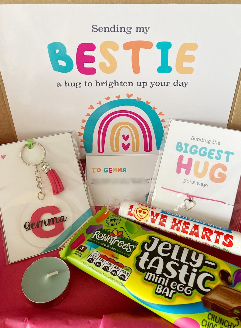 May include: A gift box with a variety of items, including a keychain with a pink tassel, a card with the name "Gemma", a candy bar, a candle, and a bracelet. The box includes a card that says "Sending my BESTIE a hug to brighten up your day".