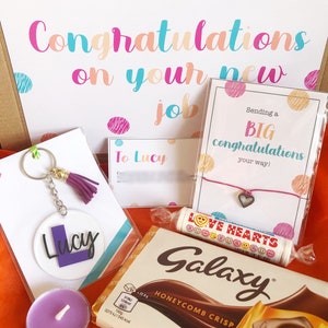 K&ouml;nnte beinhalten: Eine Geschenkbox gef&uuml;llt mit Artikeln zur Feier eines neuen Jobs. Die Box enth&auml;lt eine Karte mit der Aufschrift "Herzlichen Gl&uuml;ckwunsch zu deinem neuen Job", einen Schl&uuml;sselanh&auml;nger mit dem Namen "Lucy", eine Tafel Schokolade, eine kleine Kerze und ein Armband mit einem Herz-Anh&auml;nger.
