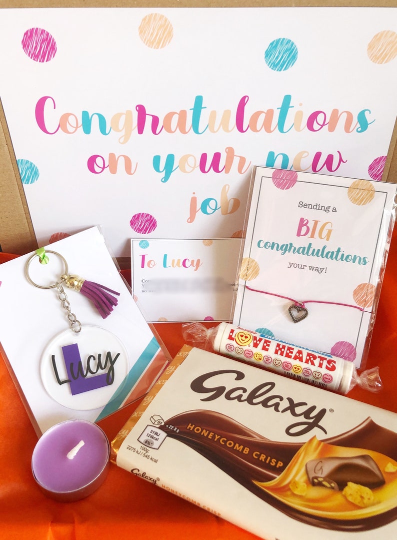 K&ouml;nnte beinhalten: Eine Geschenkbox gef&uuml;llt mit Artikeln zur Feier eines neuen Jobs. Die Box enth&auml;lt eine Karte mit der Aufschrift "Herzlichen Gl&uuml;ckwunsch zu deinem neuen Job", einen Schl&uuml;sselanh&auml;nger mit dem Namen "Lucy", eine Tafel Schokolade, eine kleine Kerze und ein Armband mit einem Herz-Anh&auml;nger.