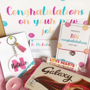 K&ouml;nnte beinhalten: Eine Geschenkbox gef&uuml;llt mit Artikeln zur Feier eines neuen Jobs. Die Box enth&auml;lt eine rosa und wei&szlig;e Karte mit dem Text "Herzlichen Gl&uuml;ckwunsch zu deinem neuen Job", eine rosa und wei&szlig;e Karte mit dem Text "An Natalie", eine rosa und wei&szlig;e Karte mit dem Text "Sende dir ein GROSSES Gl&uuml;ckwunsch auf deinem Weg!" mit einem rosa Schnurarmband mit einem silbernen Herzanh&auml;nger, einen rosa und wei&szlig;en Schl&uuml;sselanh&auml;nger mit einem klaren Kreisanh&auml;nger mit dem Text "Natalie", eine rosa Kerze und eine Galaxy Cookie Crumble Schokoladentafel.