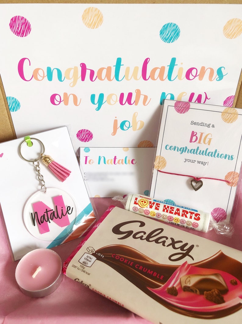 K&ouml;nnte beinhalten: Eine Geschenkbox gef&uuml;llt mit Artikeln zur Feier eines neuen Jobs. Die Box enth&auml;lt eine rosa und wei&szlig;e Karte mit dem Text "Herzlichen Gl&uuml;ckwunsch zu deinem neuen Job", eine rosa und wei&szlig;e Karte mit dem Text "An Natalie", eine rosa und wei&szlig;e Karte mit dem Text "Sende dir ein GROSSES Gl&uuml;ckwunsch auf deinem Weg!" mit einem rosa Schnurarmband mit einem silbernen Herzanh&auml;nger, einen rosa und wei&szlig;en Schl&uuml;sselanh&auml;nger mit einem klaren Kreisanh&auml;nger mit dem Text "Natalie", eine rosa Kerze und eine Galaxy Cookie Crumble Schokoladentafel.